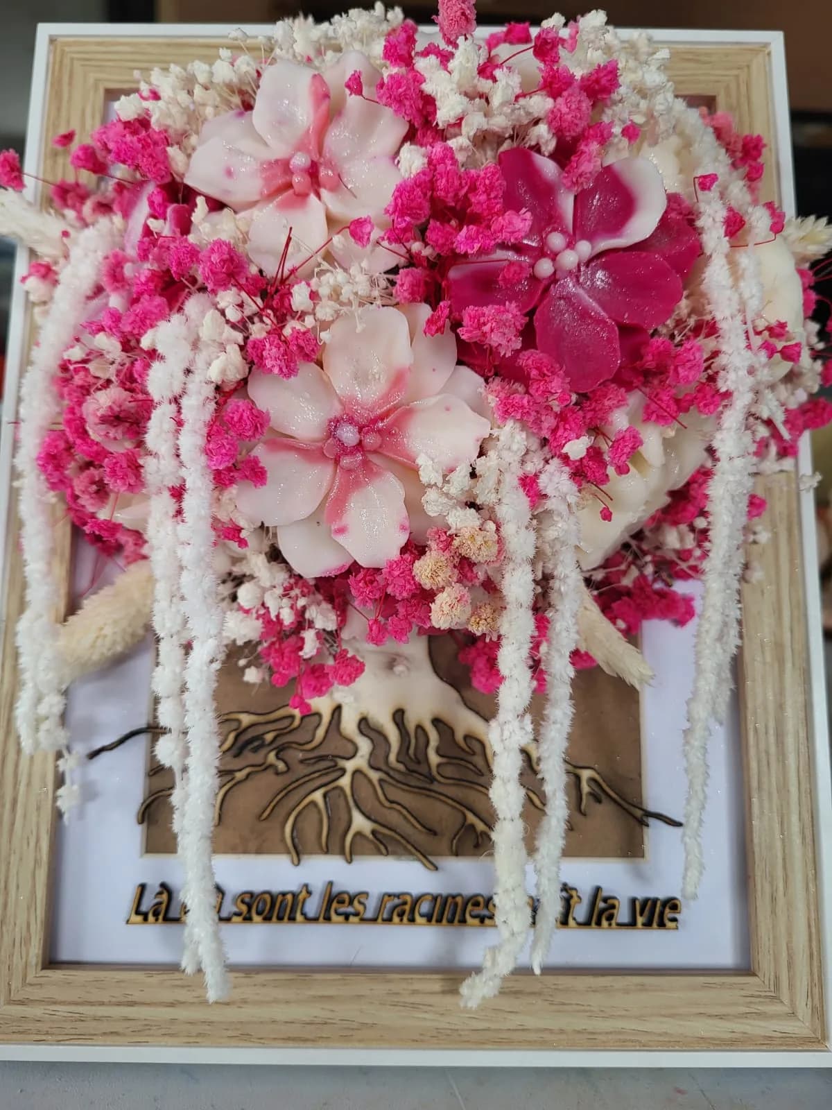 Cadre en bois clair : bouquet de fleurs de cire rose vif et blanches au-dessus d’un arbre de vie en bois découpé